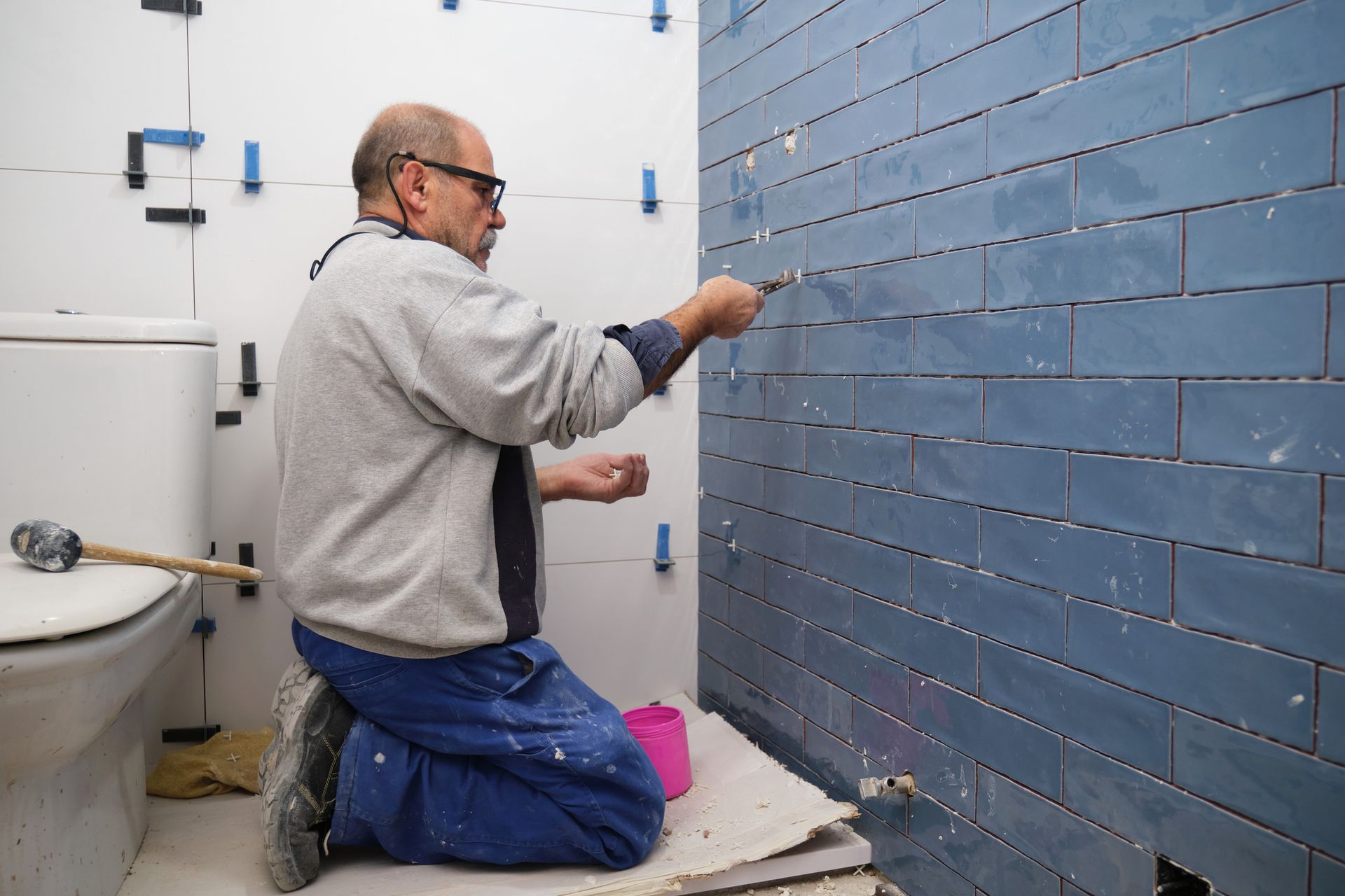 Man kneeling, applying grout to blue tiles on bathroom wall.