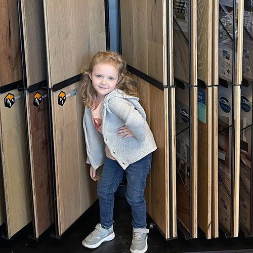 Girl in jeans and gray sweater poses in front of wood flooring samples.