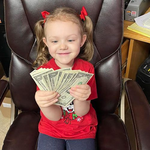 Young child with pigtails holding several dollar bills, smiling in a chair.