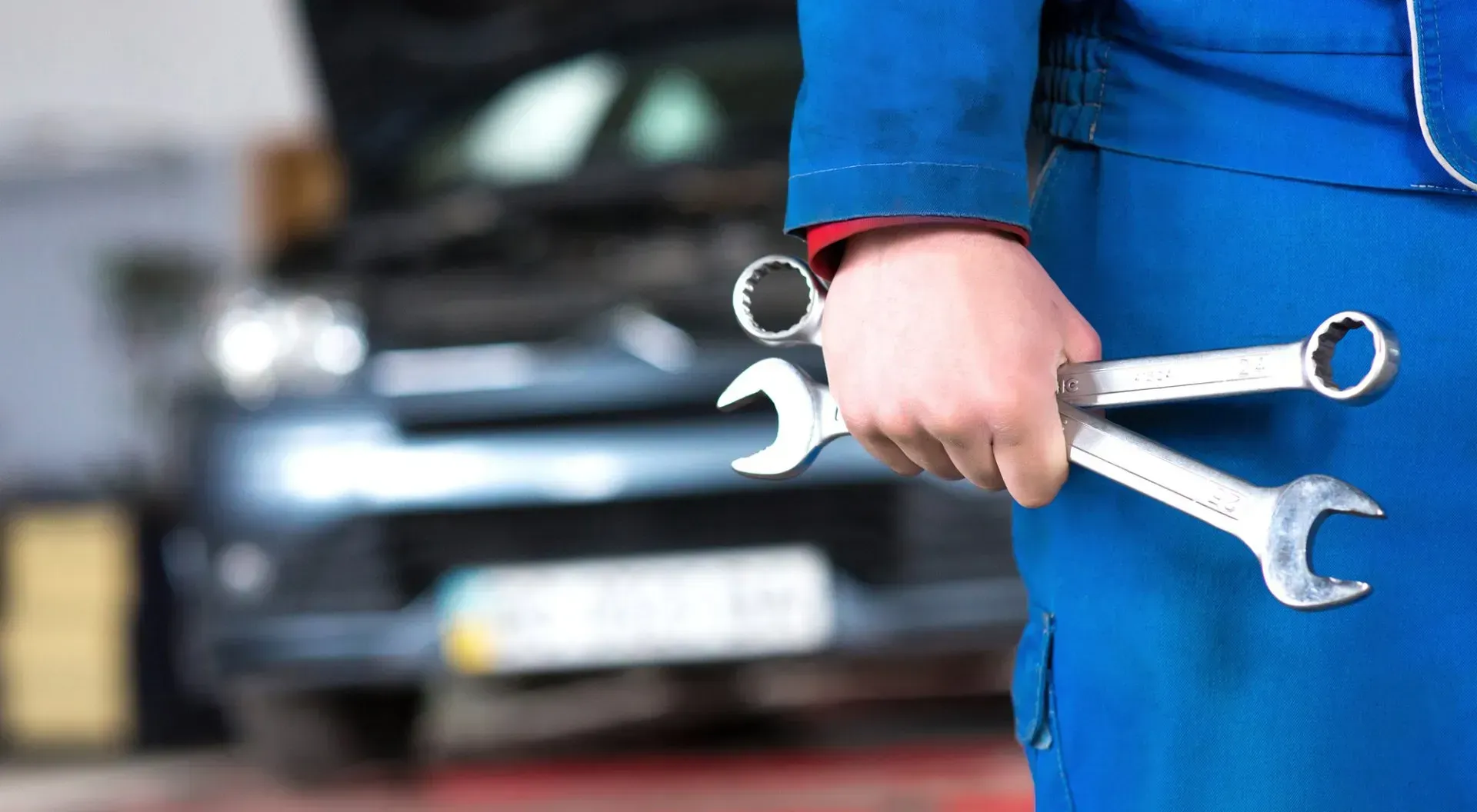 Mechanic in blue overalls holding wrenches, car in background.