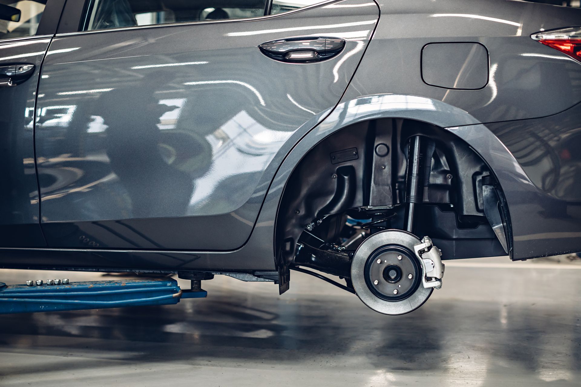 Grey car, rear wheel removed, in a repair shop. Brake system visible.