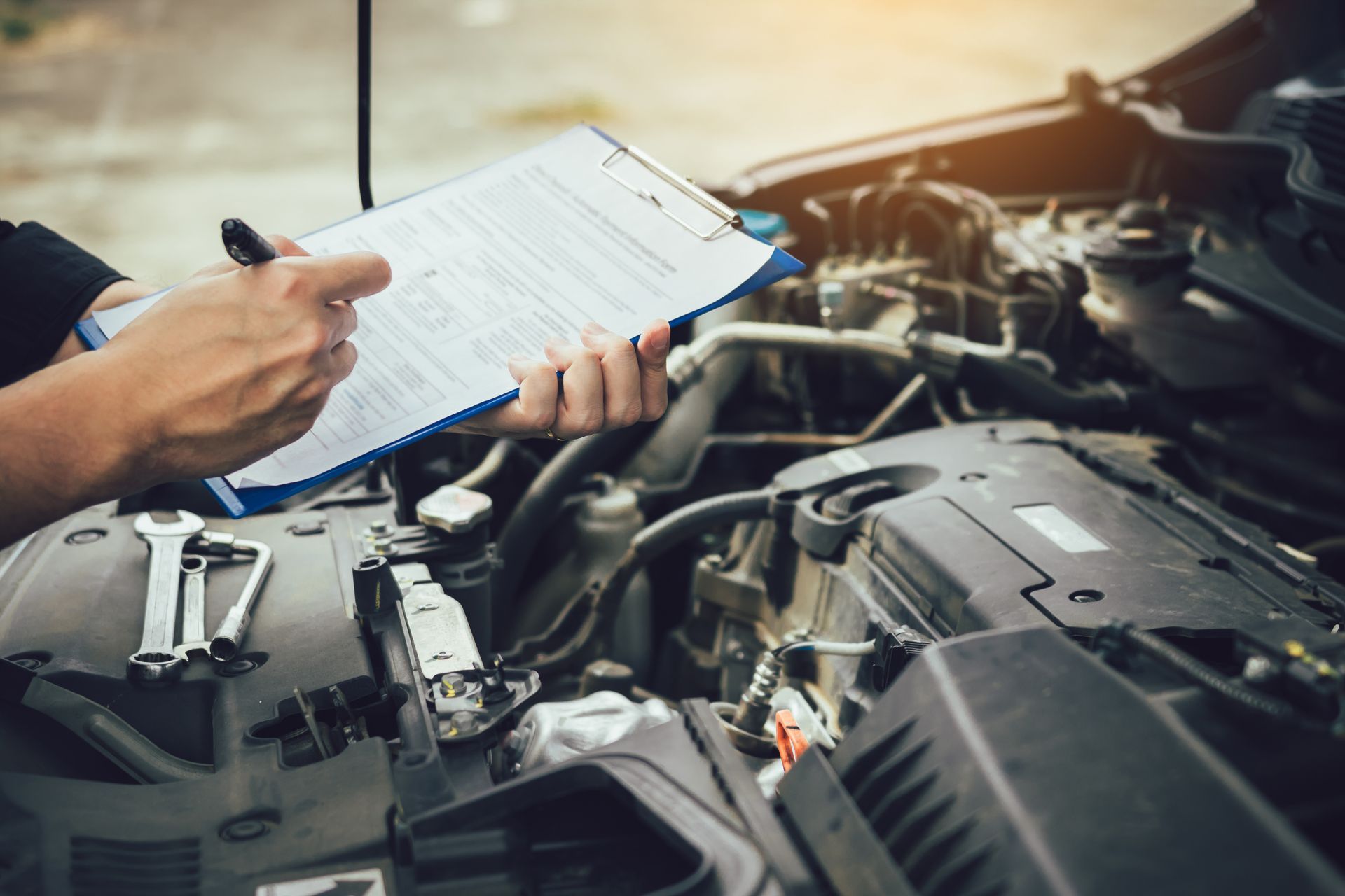 Mechanic inspecting car engine, holding clipboard and pen. Tools visible, outdoors.