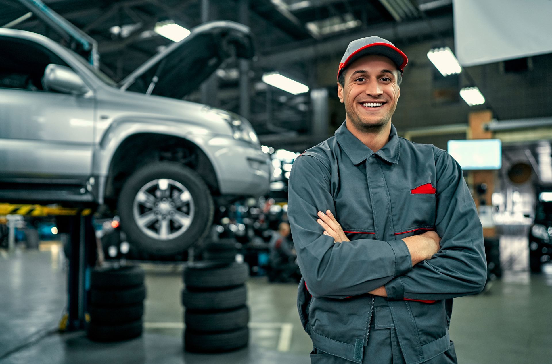 Smiling mechanic in gray jumpsuit, arms crossed, in auto repair shop with lifted car.