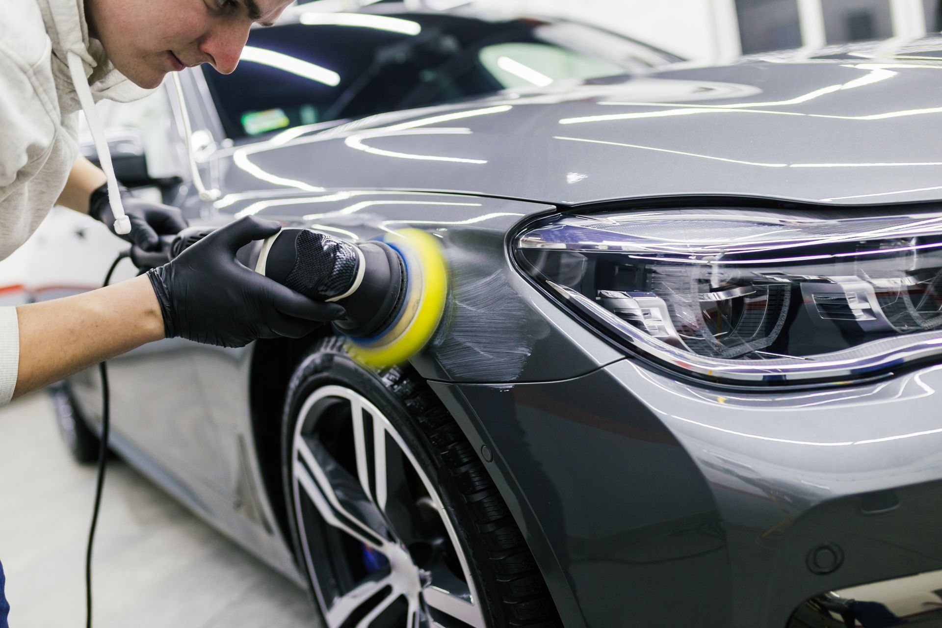 Person polishing a gray car with a buffer, black gloves, bright yellow pad.