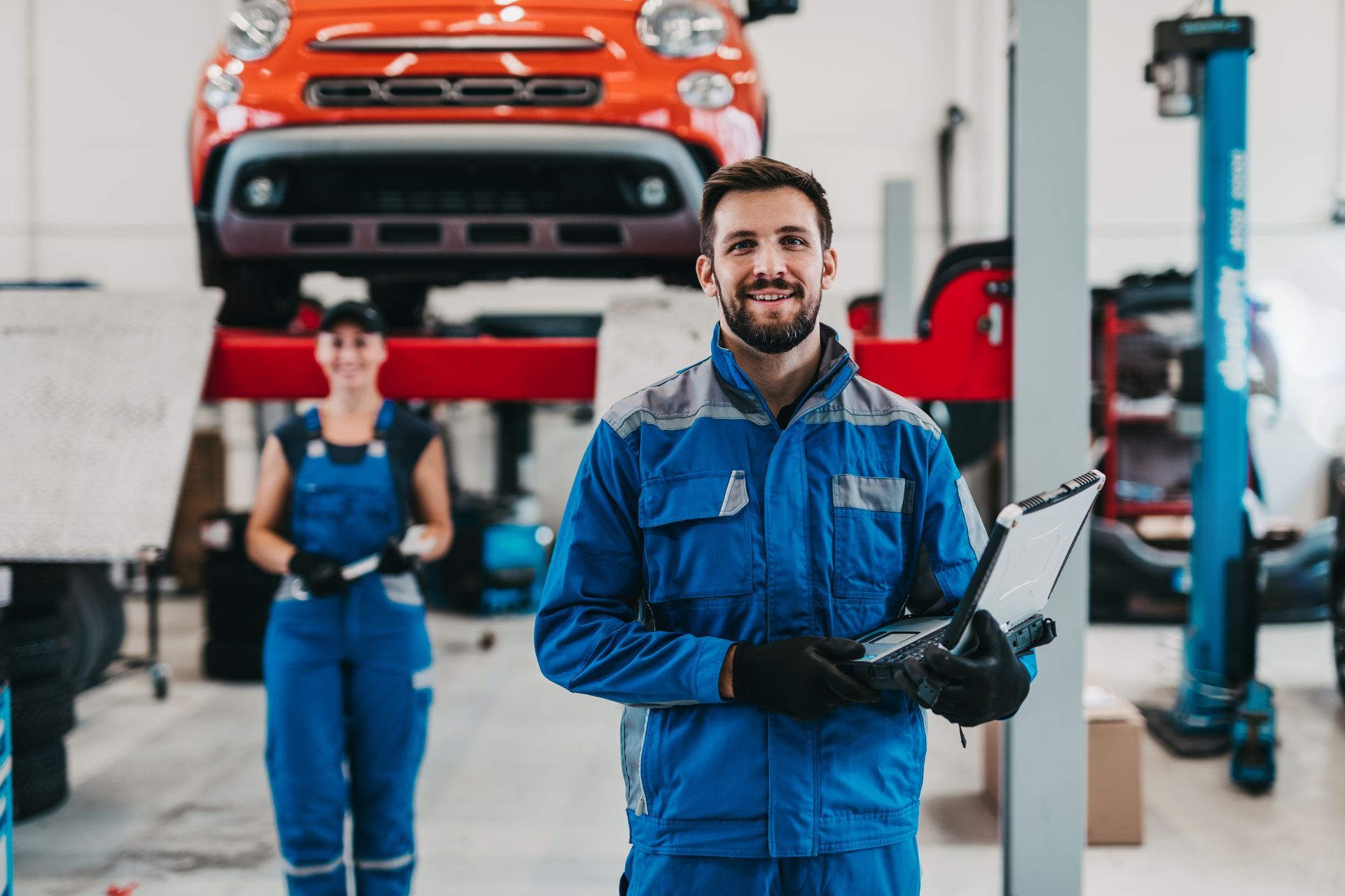 Mechanic in blue overalls smiles, holding clipboard in a car repair shop. Another mechanic in the background. Orange car on a lift.