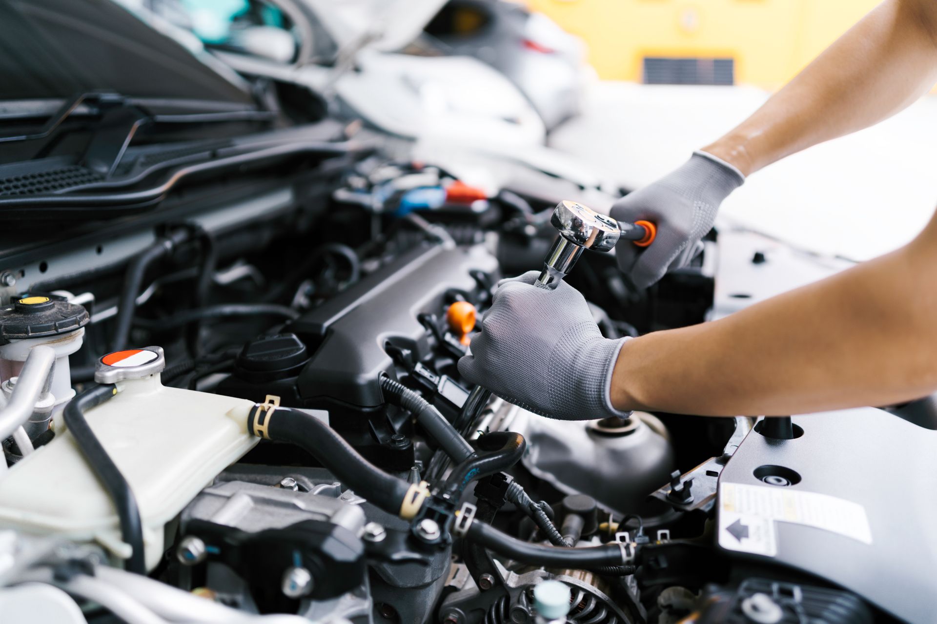 Mechanic working on a car engine with a wrench, wearing gloves in a garage.