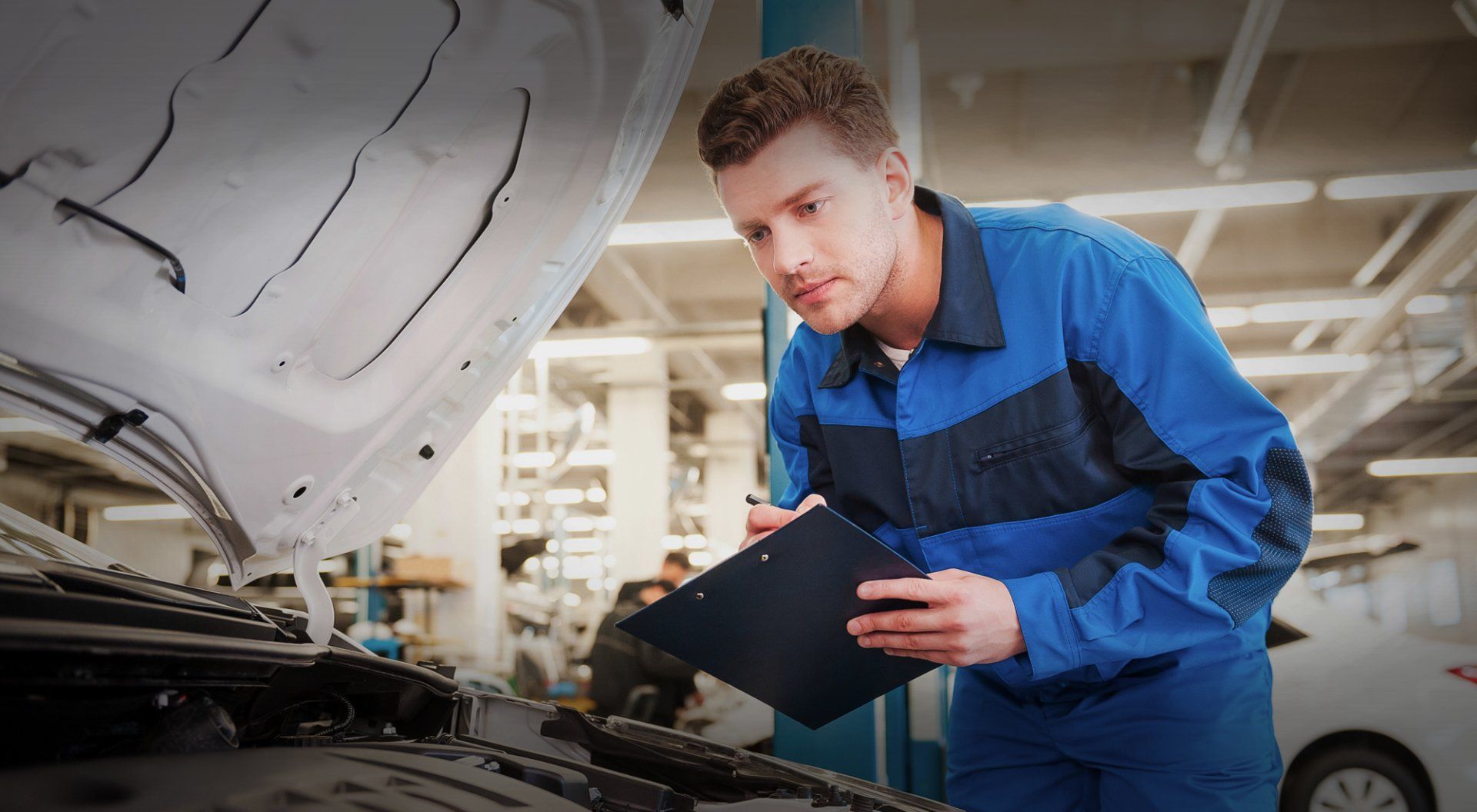 Mechanic in blue coveralls examines car engine, holding clipboard, in a garage.