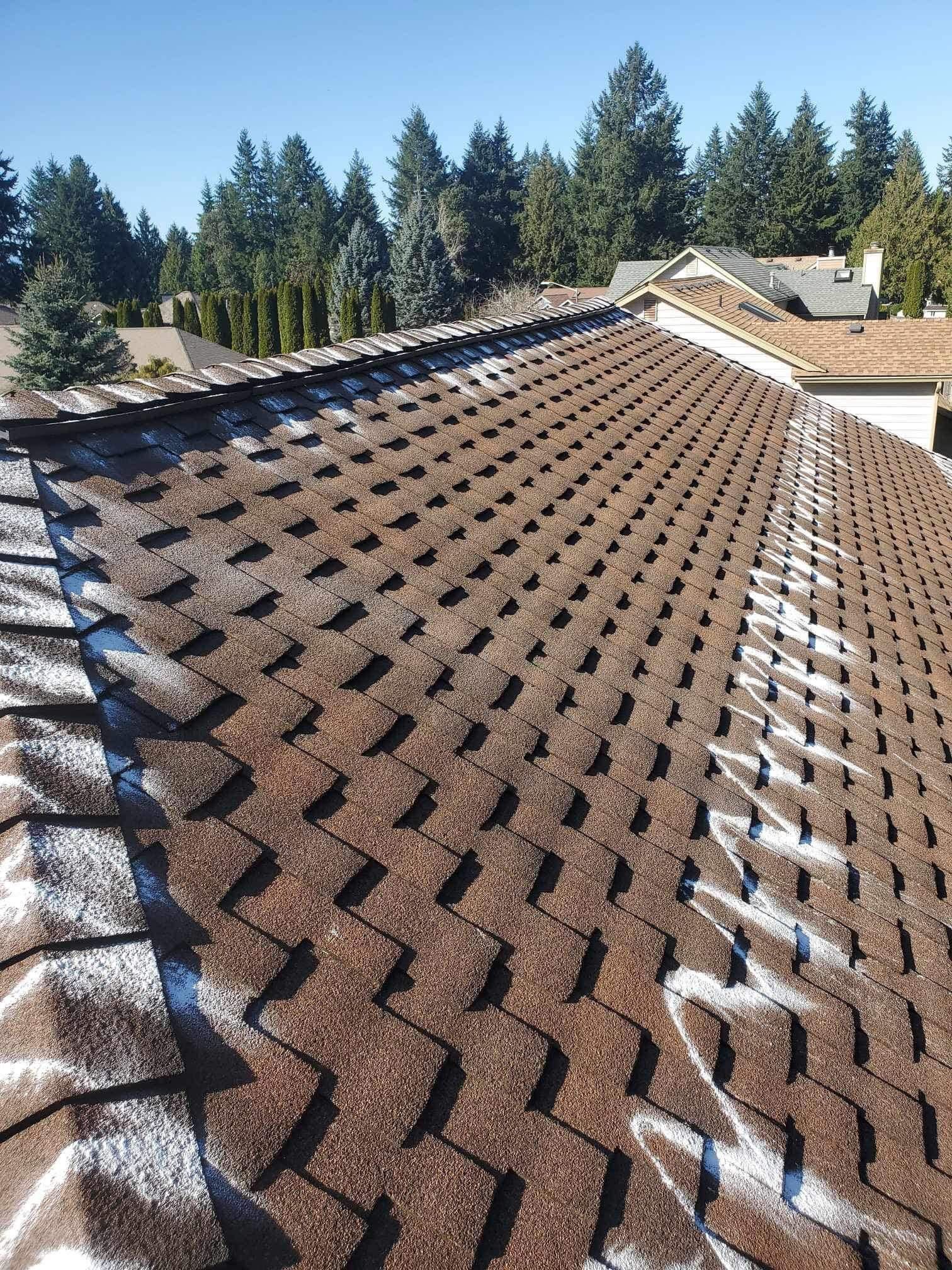 Brown asphalt shingle roof with patches of snow under a bright blue sky, with trees in the background.