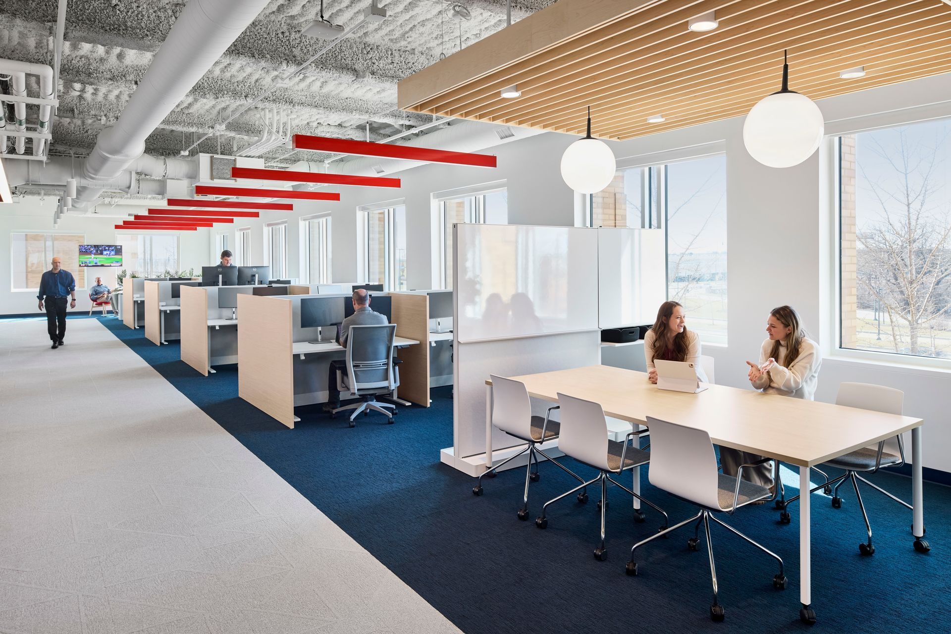 Modern office interior: rows of workstations, meeting table, large windows, people working, blue and white carpet, red accents.