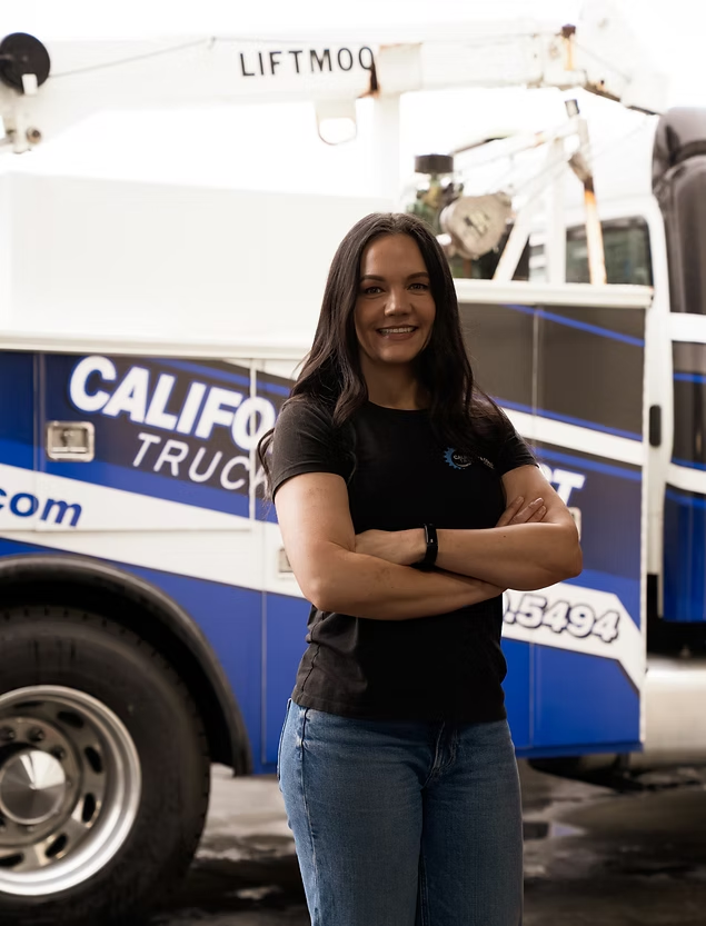 A person with long dark hair stands with arms crossed in front of a blue and white California truck. | Kensington Motor Cars