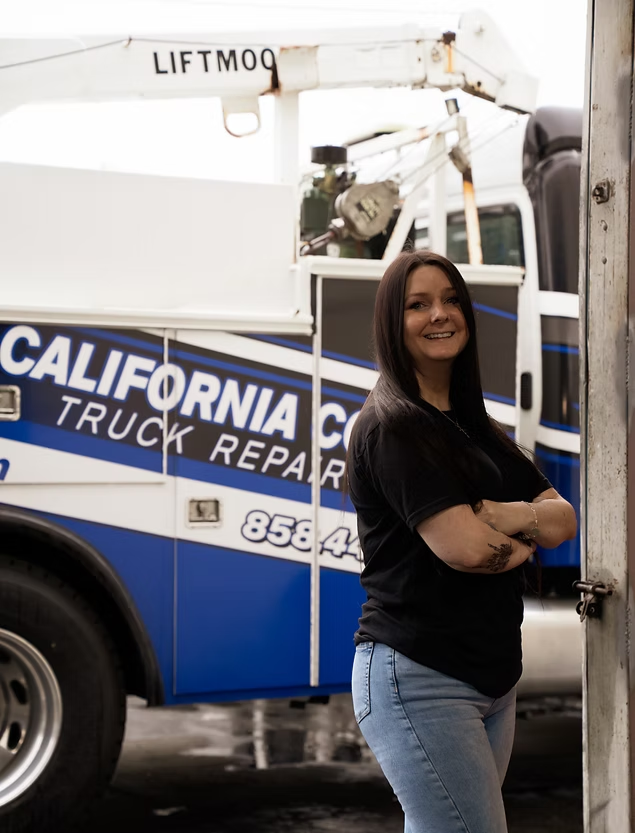 A person with long dark hair and crossed arms standing in front of a blue and white California Truck Repair utility truck. | Kensington Motor Cars