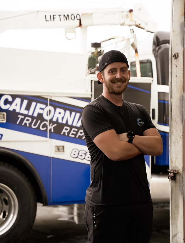 A smiling person with arms crossed, wearing a black t-shirt and cap, standing in front of a California Truck Repair vehicle. | Kensington Motor Cars