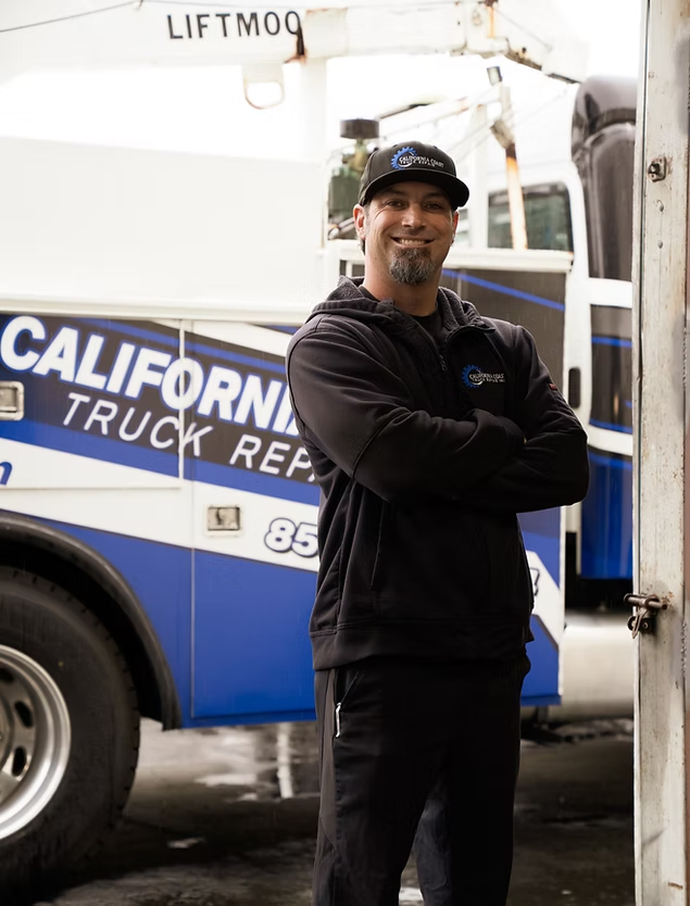 A smiling person with a goatee wearing a black hooded sweatshirt and baseball cap, standing before a service truck. | Kensington Motor Cars