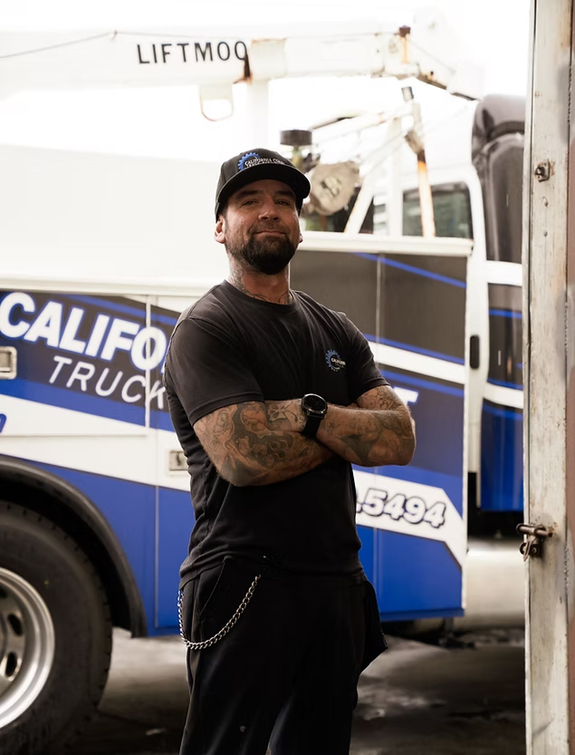 A person with tattooed arms stands with crossed arms in front of a blue and white tow truck. | Kensington Motor Cars