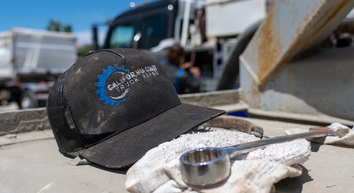 A black hat with a blue logo sits on a truck bed next to a work glove and a wrench, with a mechanic working in the background. | California Coast Truck Repair