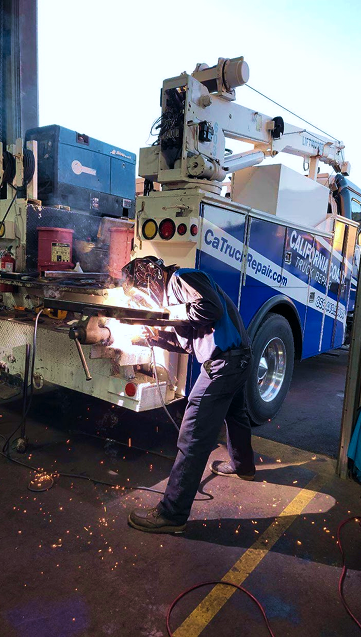 Person welding on a truck bed, sparks flying.  Blue and white truck, crane in the background, outdoor setting. | California Coast Truck Repair