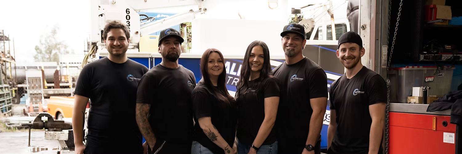 Six people in matching black shirts stand in a row in front of a service vehicle at an industrial site. | Kensington Motor Cars