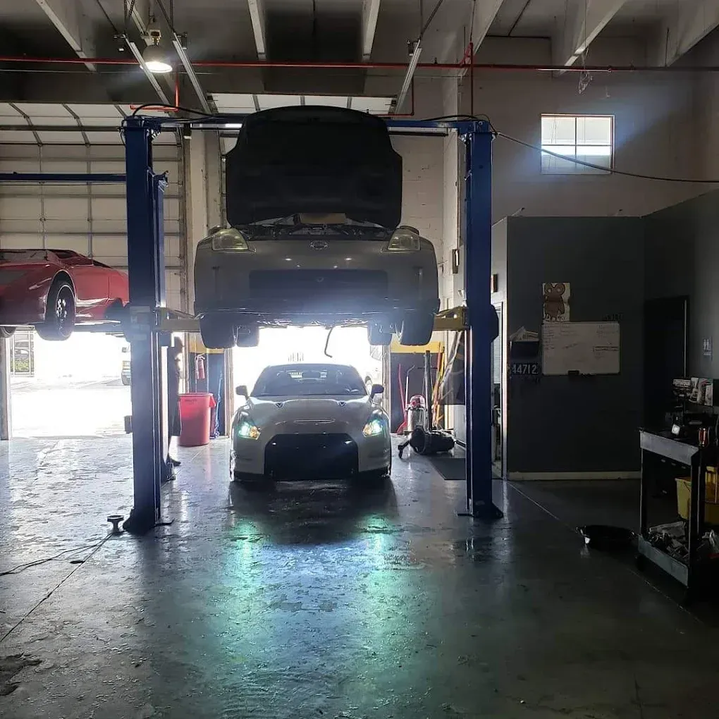 A car repair shop with a car on a lift, a white car in front, and a red car on another lift in the background.