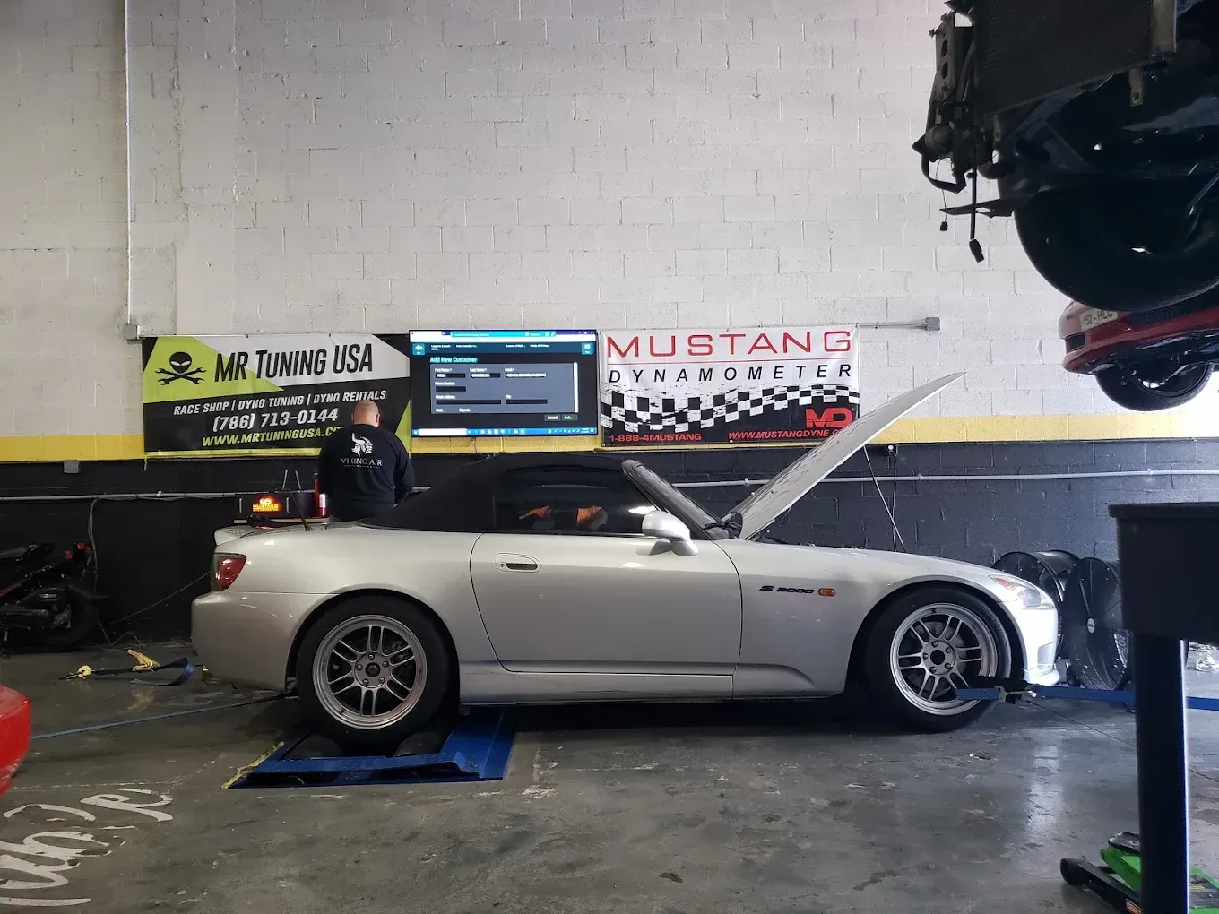 Silver Honda S2000 roadster in a garage with its hood open. A mechanic is working near the car, with a dyno setup visible.