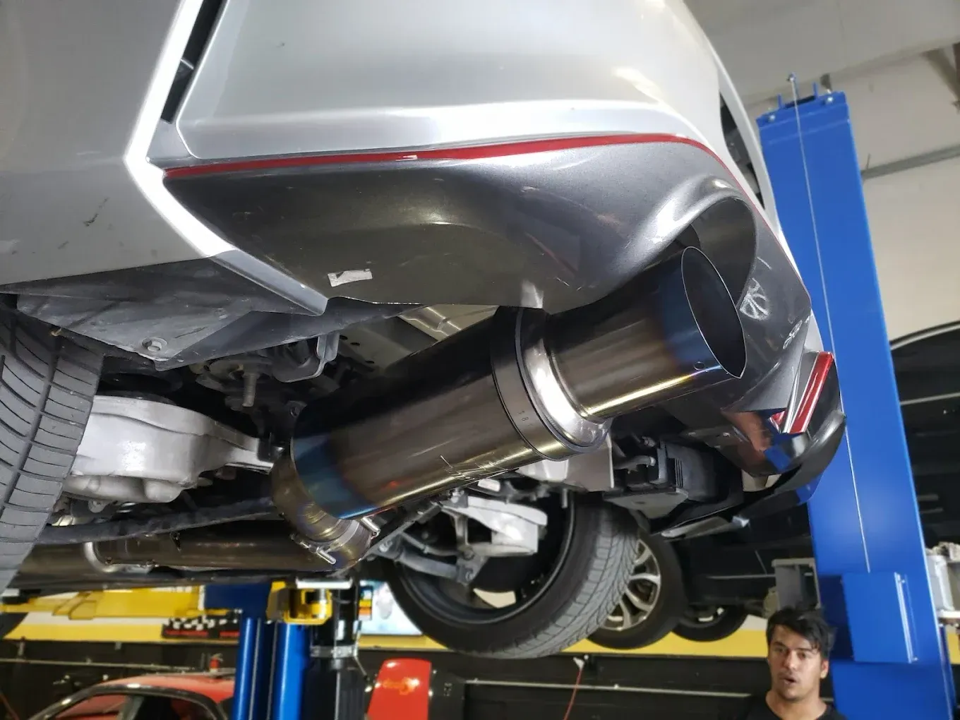 A car's underside, showcasing a large, blue-tipped exhaust system installed in a garage, with a man looking at the camera.
