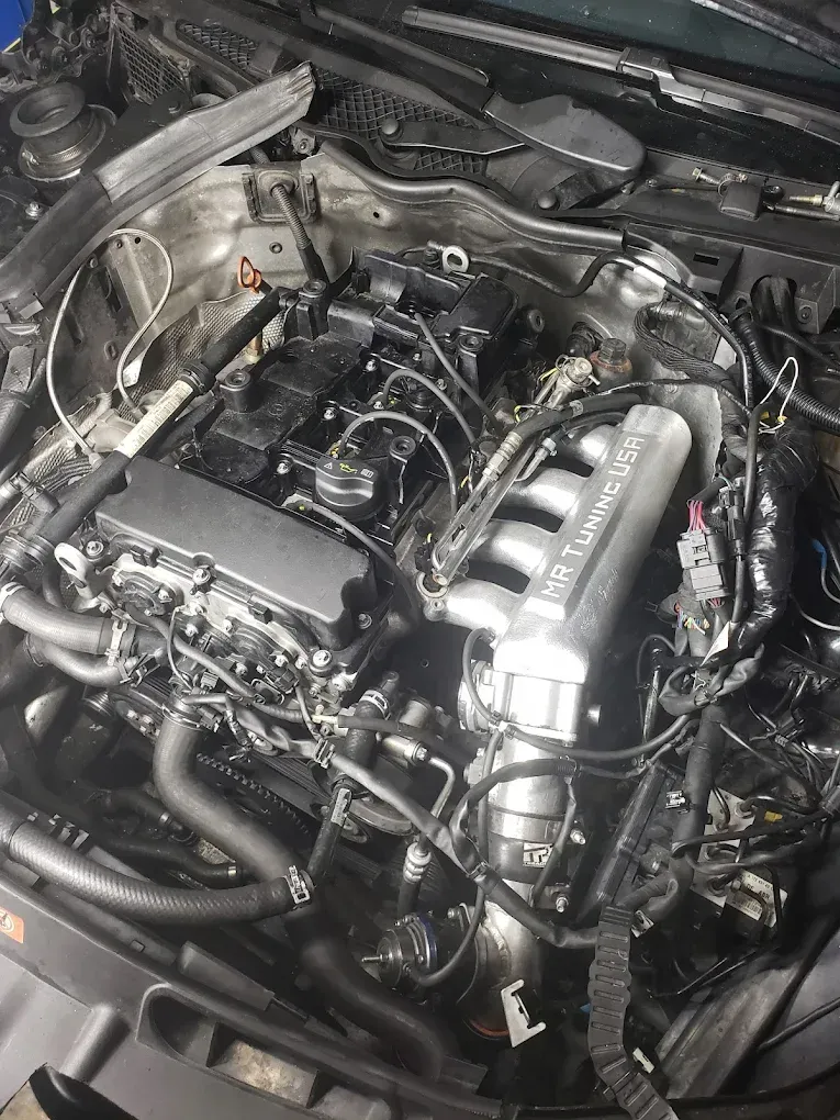 Engine bay of a car, top-down view. Engine is partially disassembled, showing components like the intake manifold and various hoses.