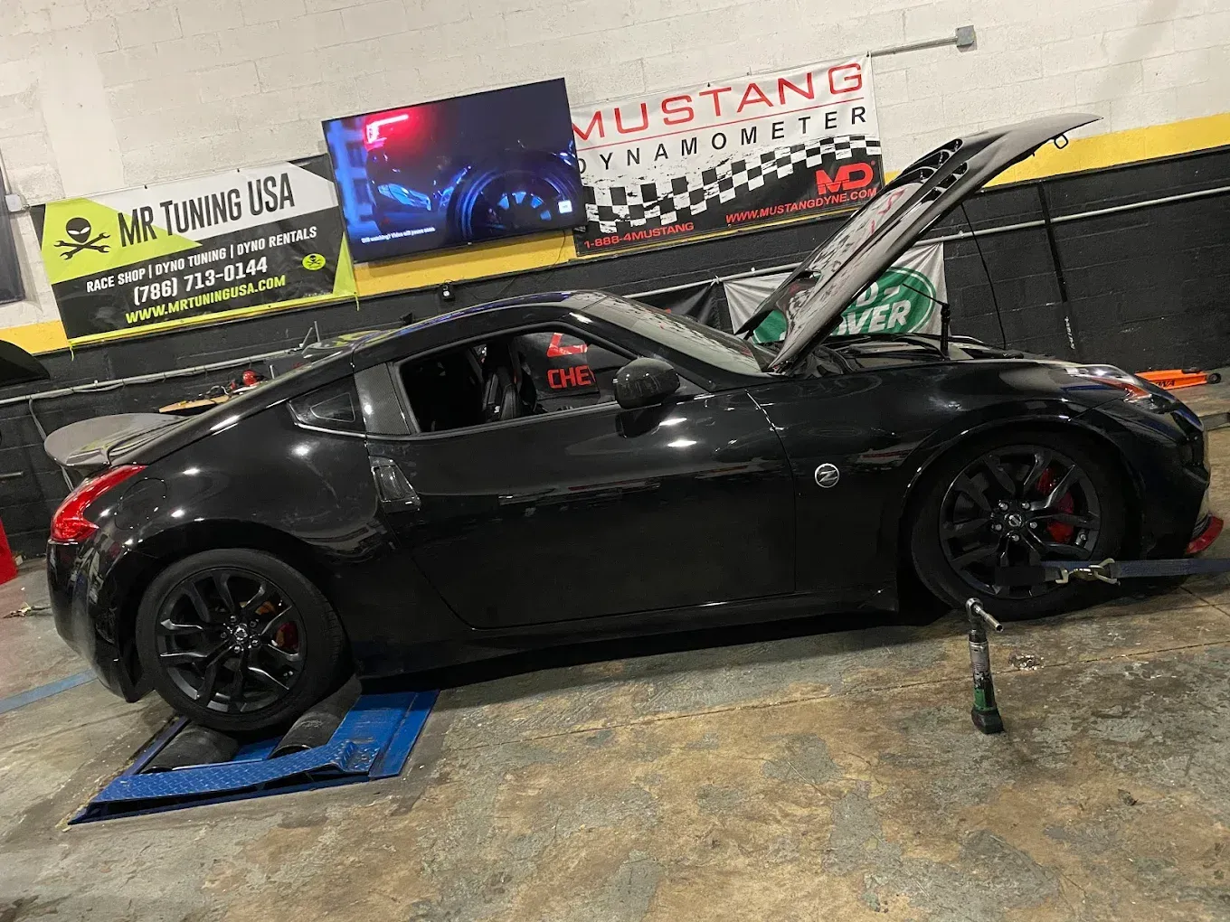 Black Nissan 370Z on a dynamometer with hood open in a shop. Red brake calipers and a spoiler are visible.
