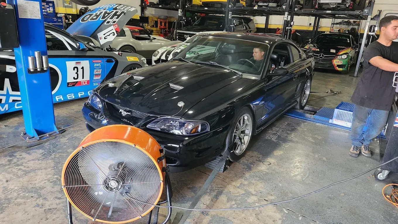 Black Ford Mustang on a dynamometer in a garage, with a large orange fan in front and a man inside.