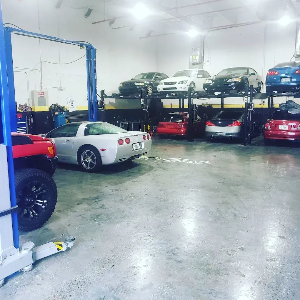 Inside a car repair shop, a silver Corvette and several other cars are on lifts. The shop has lifts and bright lighting.