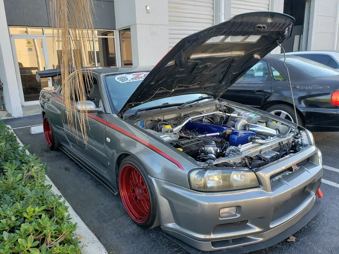 Gray custom sports car with red wheels, hood open, parked outdoors near a building.