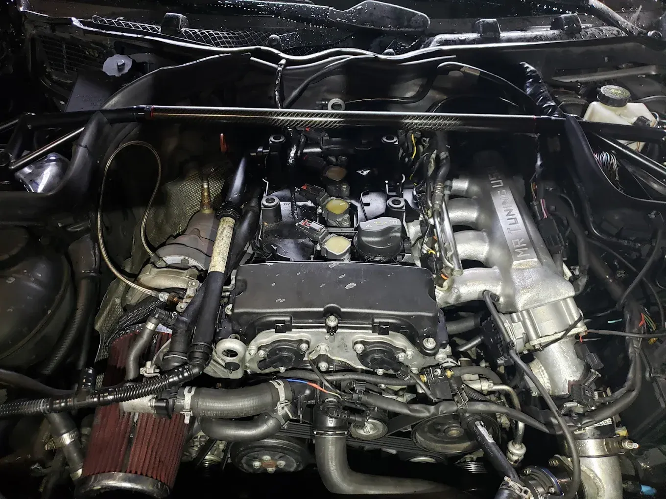 Engine bay of a black car with visible engine components, including a red air filter, silver brace, and black engine cover.