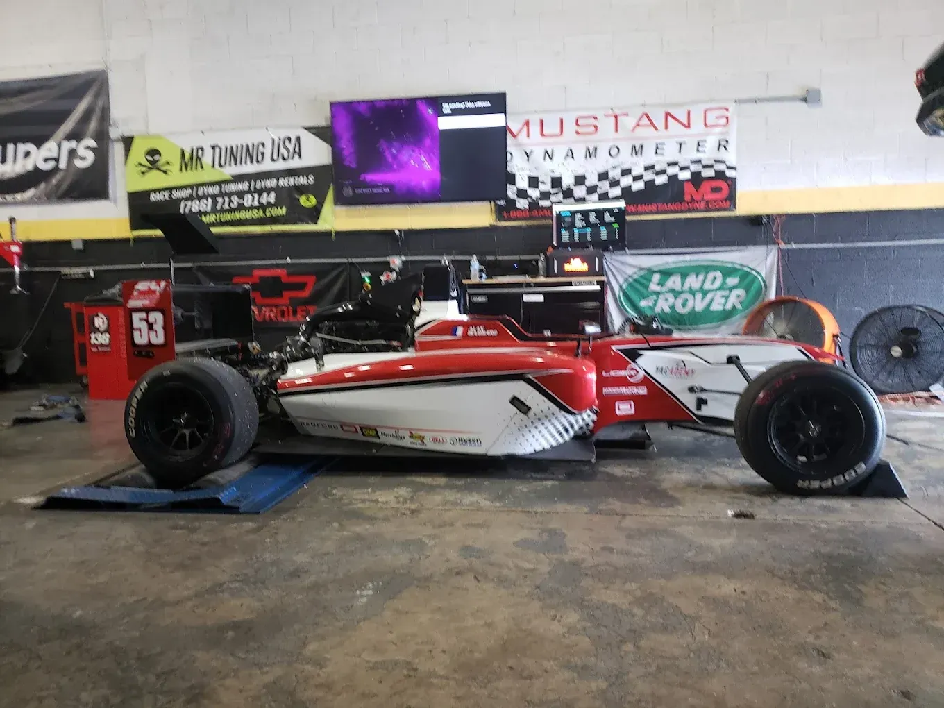 A red and white race car on a dynamometer in a garage, with black wheels and sponsor logos on the walls.