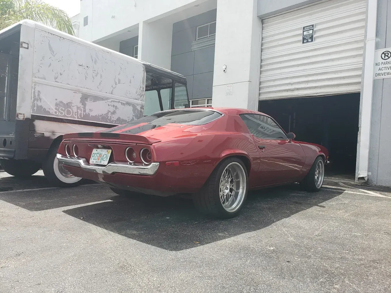 Red classic car parked outside a garage. The car is low to the ground, and a truck is next to it.