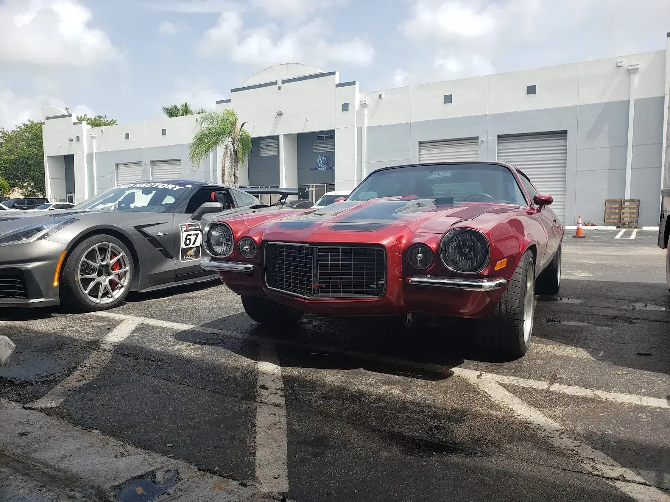 A classic red Camaro parked next to a gray Corvette in a parking lot in front of a light-colored building.