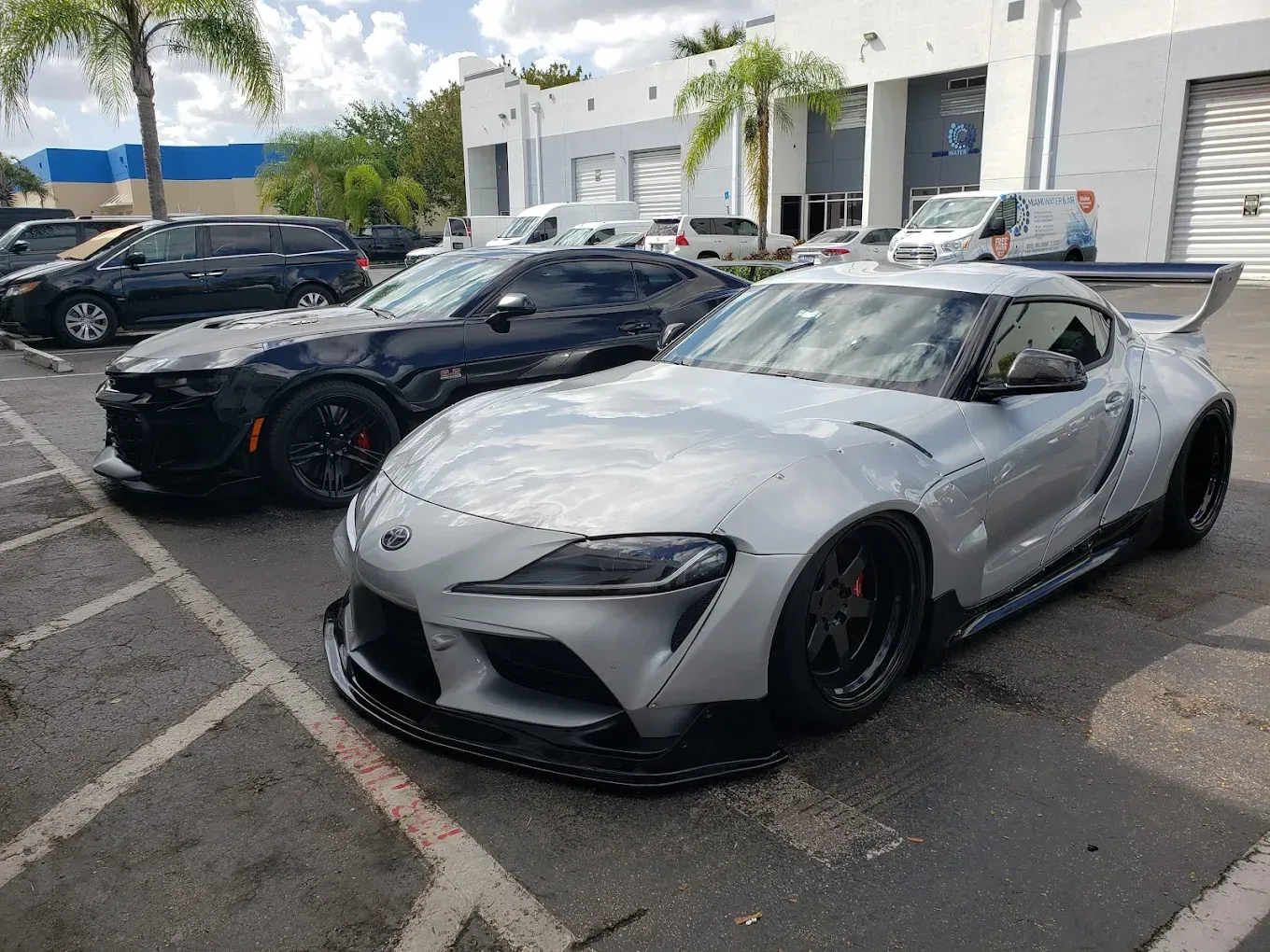 Silver Toyota Supra modified with a wide body kit, parked next to a black Camaro in a parking lot.