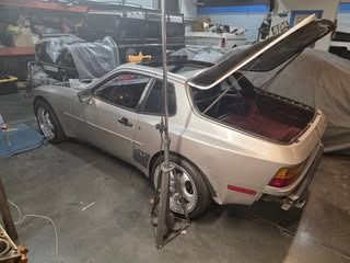 Silver Porsche 944 with open hatch, in a garage with tools and covered cars.
