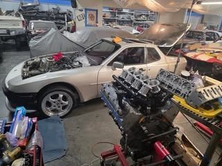 White Porsche 944 in a garage with an engine. Open hood, other car under cover, engine parts visible.