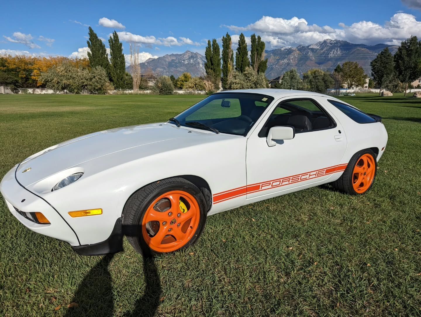 White Porsche 928 with orange wheels and side stripe parked on grass with mountains in the background.