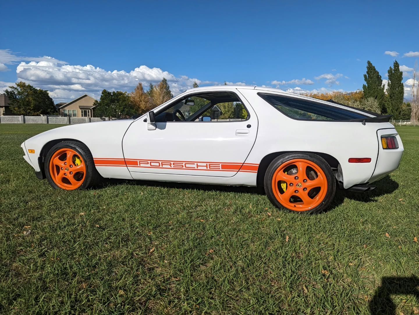 White Porsche 928 sports car with orange wheels and side stripe parked on grass under a blue sky.