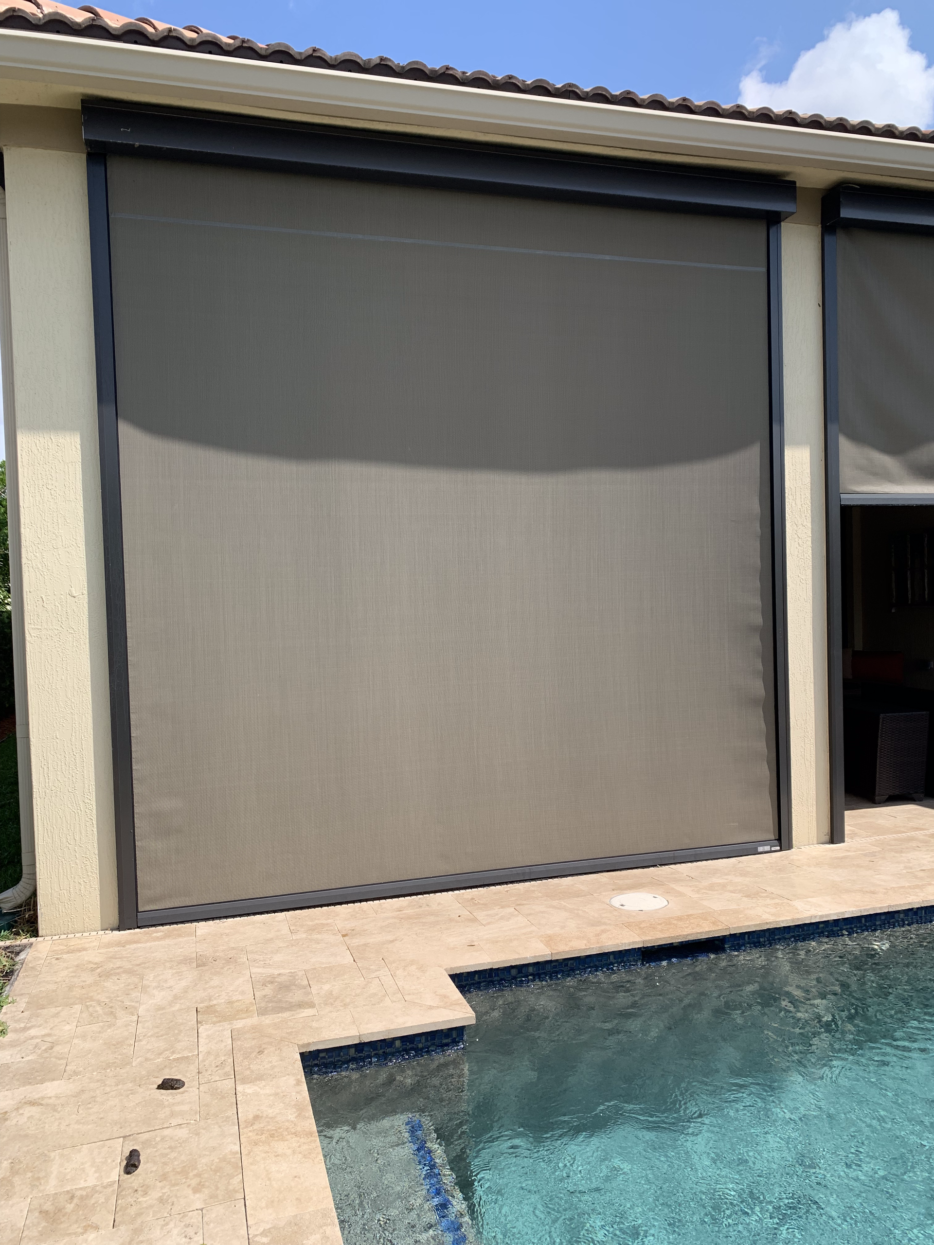 Large, rolled-up shade covers the outdoor pool patio. Tan fabric, black trim. Blue pool water and tile.