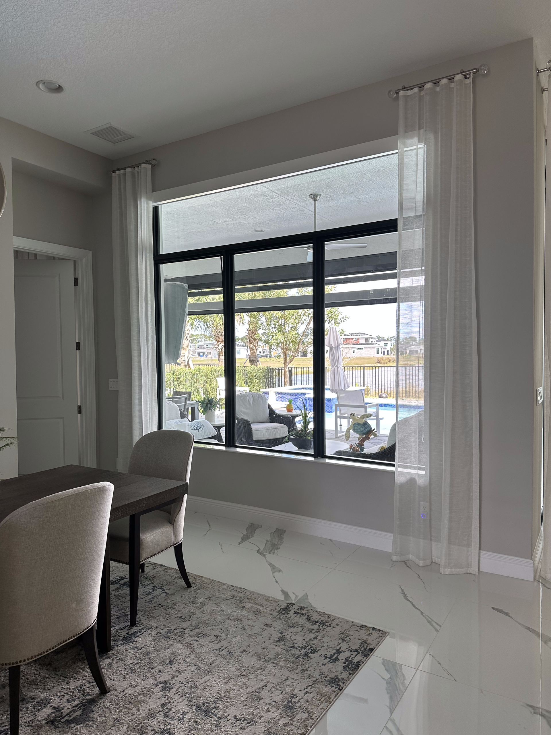 Dining room with large window, sheer white curtains, overlooking a pool area. Marble floor and neutral furnishings.