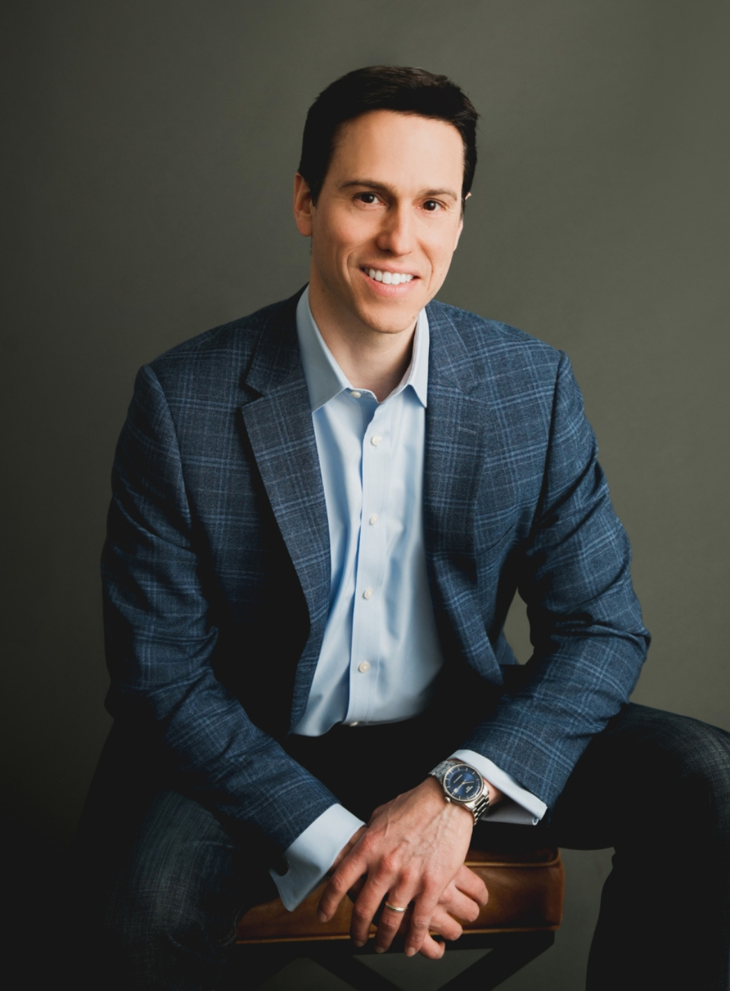 Man in blue blazer and open light blue shirt, seated with hands clasped, smiling, against a gray background.