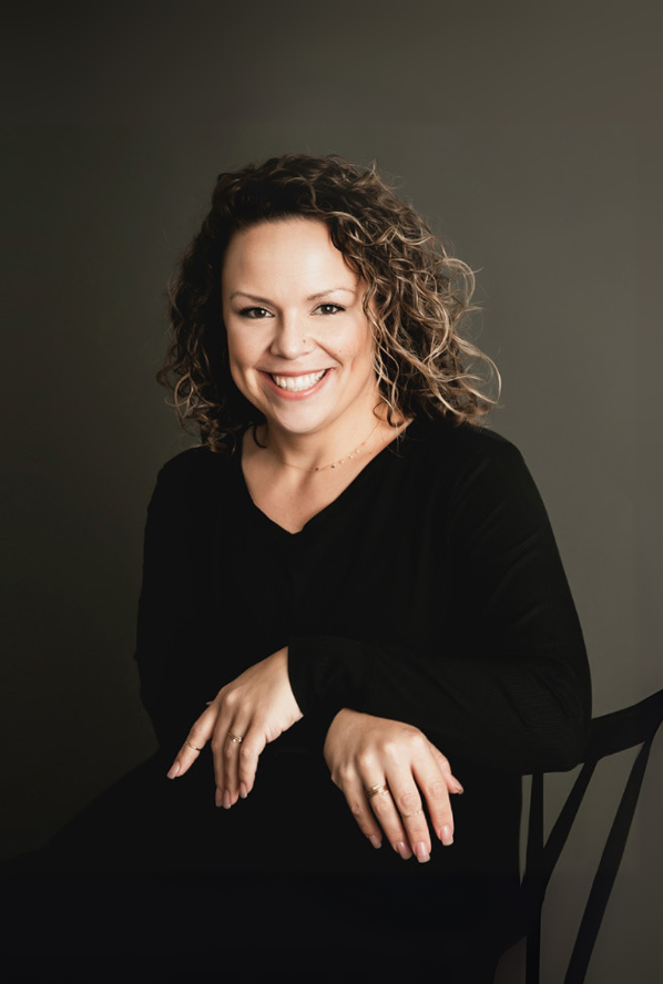 Woman with curly hair, smiling, wearing a black sweater, seated against a grey background.