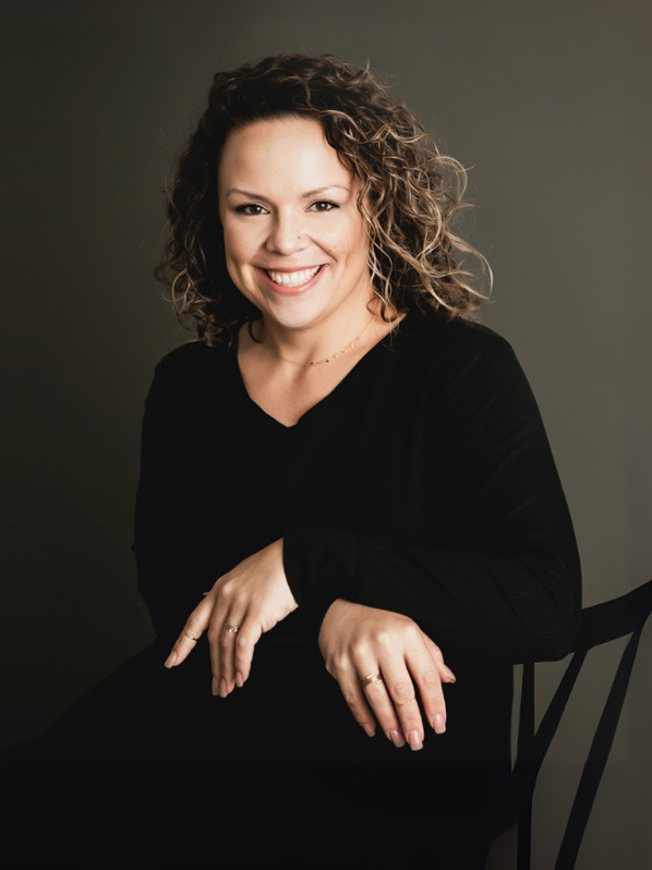 Woman with curly hair smiles, seated, wearing black, on a chair against a gray background.