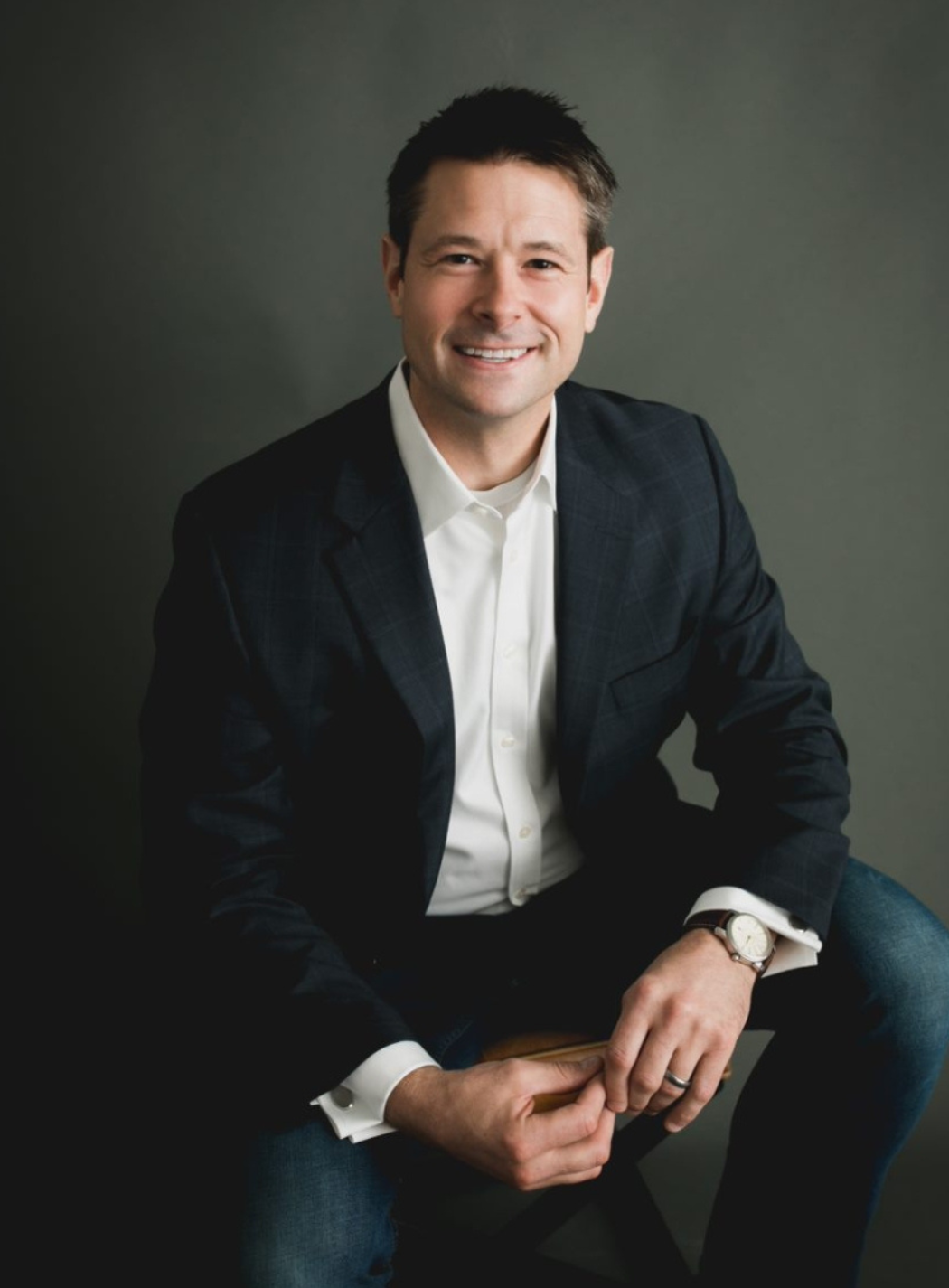 Man in a suit jacket and jeans, smiling, seated with hands clasped, gray background.