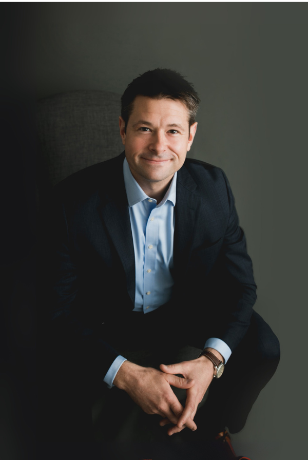 Man in a suit and blue shirt, smiling, sitting with hands clasped, dark background.