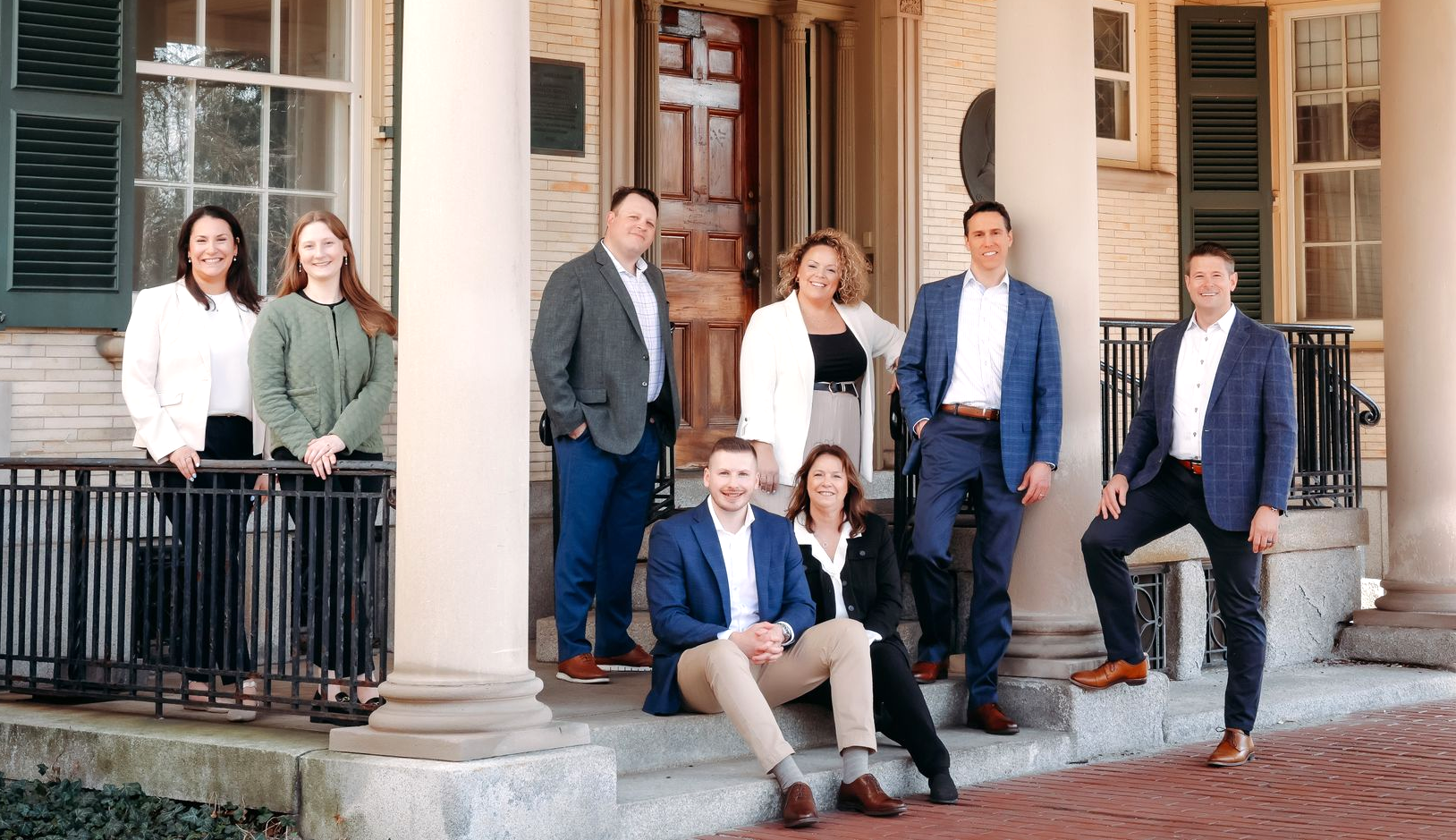 Group of eight people posing on the steps of a brick building with columns and large windows