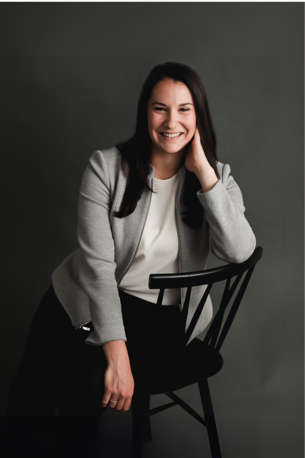 Woman in blazer smiles, sitting on chair against a gray backdrop.