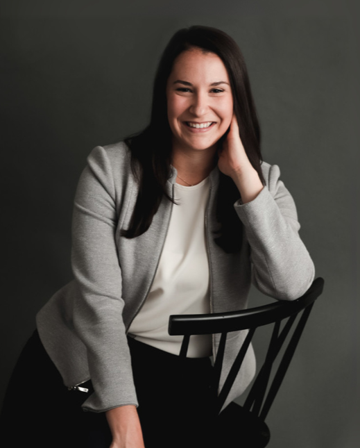 Woman in business attire smiles, sitting in a chair, hand near face, against a grey background.