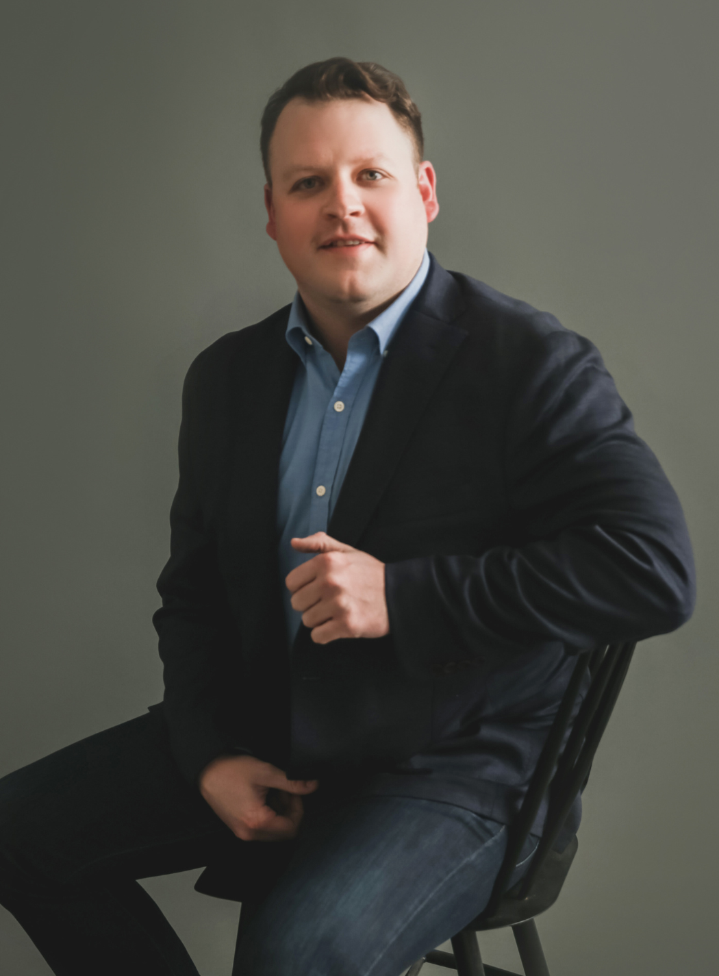 Man in blazer and blue shirt seated on a chair, looking towards the camera, neutral expression.