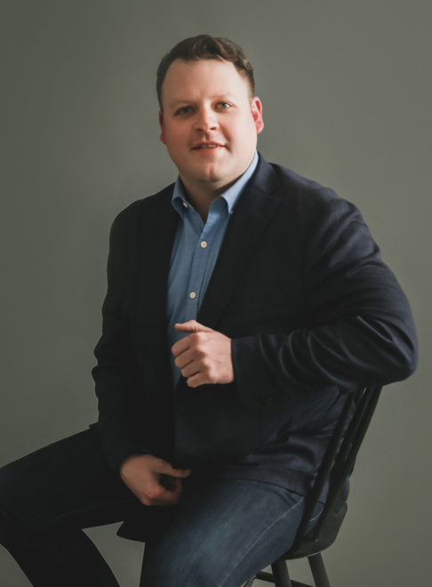 Man in blazer and blue shirt seated on a chair, looking towards the camera, neutral expression.